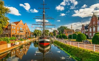 Museumssegelschiff mit hohen Masten liegt in einem Kanal in Papenburg, flankiert von traditionellen Backsteingebäuden und gepflegten Gärten unter blauem Himmel.