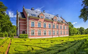 Palsjö Schloss, ein rosa Barockschloss mit grauem Dach, umgeben von ornamentalem Heckenlabyrinth unter blauem Himmel bei Helsingborg.