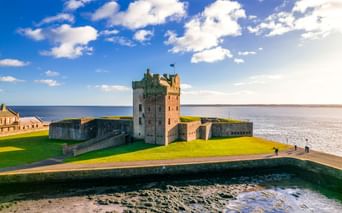Broughty Castle in Dundee mit Steinturm und Befestigungen auf grüner Wiese neben Flussmündung unter blauem Himmel mit weißen Wolken.