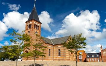 Backsteinkirche Erlöserkirche in Esbjerg mit hohem Spitzturm und Rundbogenfenstern, umgeben von jungen Bäumen auf einem gepflasterten Platz.