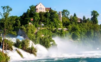 Rheinfall in Schaffhausen mit kraftvoll strömendem Wasser, Gischt und Regenbogen. Schloss Laufen thront auf grünem Hügel mit Schweizer Flagge.