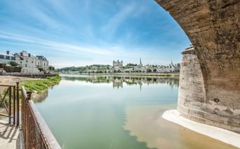 Blick unter einem Steinbrückenbogen auf die Loire in Saumur, mit historischen Gebäuden und Kirchtürmen, die sich im ruhigen Wasser spiegeln.