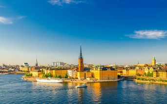Panoramablick auf Stockholms Gamla Stan mit bunten historischen Gebäuden am Wasser und Kirchtürmen unter blauem Himmel.