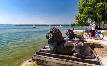 Zwei Radfahrer mit Rädern neben bronzenen Löwenstatuen am Starnberger See. Segelboote auf dem See, Berge im Hintergrund unter blauem Himmel.