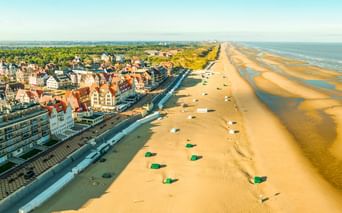 Luftaufnahme der Küstenstadt De Haan in Belgien mit traditioneller Architektur, breitem Sandstrand und Nordseeküste in der Ferne.