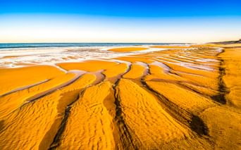 Breiter Sandstrand in Mimizan Plage mit gewellten goldenen Sandmustern und flachen Wasserpfützen. Der Atlantik trifft auf klaren blauen Himmel.