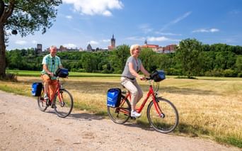 Zwei Radfahrer mit blauen Packtaschen auf einem Weg mit der historischen Skyline von Bad Wimpfen im Hintergrund mit Kirchtürmen und mittelalterlichen Gebäuden.