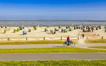 Radfahrer auf Weg entlang Neuharlingersiel Strand mit bunten Strandkörben im Sand, Nordseeküste im Hintergrund unter blauem Himmel.
