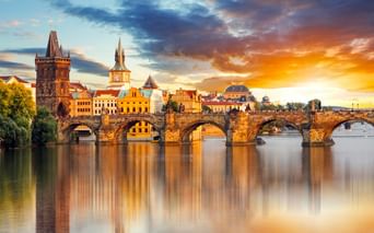 Karlsbrücke über die Moldau in Prag bei Sonnenuntergang mit historischen Türmen und Gebäuden, die sich im ruhigen Wasser unter dramatischem Himmel spiegeln.