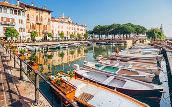 Hafen in Desenzano mit vertäuten Booten entlang der Promenade. Historische Gebäude säumen das Ufer, Bäume und eine Brücke im Hintergrund.