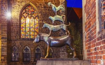 Bronze statue of Bremen Town Musicians showing stacked animals (donkey, dog, cat, rooster) illuminated at night beside a Gothic church with stained glass.