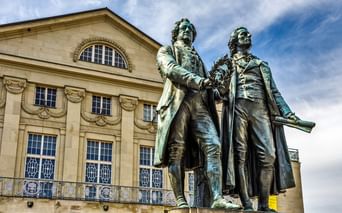 Bronzestatue zweier Männer in Kleidung des 18. Jahrhunderts auf einem Sockel, vor einem neoklassizistischen Gebäude mit Bogenfenstern.