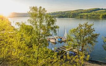 Blick auf den Baldeneysee mit Segelbooten an einem Holzsteg. Grüne Bäume rahmen die Szene, sanfte Hügel im Hintergrund unter sonnigem Himmel.