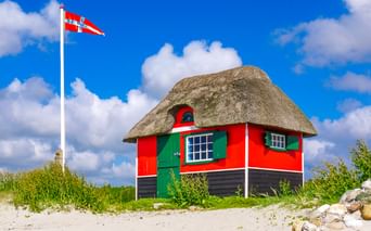 Rot-schwarzes Reetdach-Strandhäuschen mit grünen Fensterläden auf Sanddüne. Dänische Flagge an weißem Mast vor blauem Himmel auf Ærø.