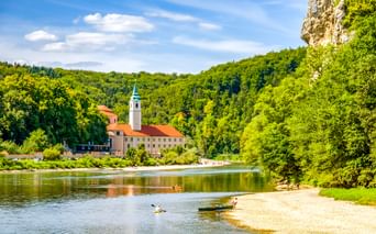 Weltenburg Abbey with bell tower beside the Danube River, surrounded by green forested hills. Kayakers paddle near a sandy beach in the foreground.