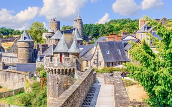Burg Fougères mit Steintürmen und befestigten Mauern, umgeben von historischen Gebäuden und grüner Vegetation unter blauem Himmel mit weißen Wolken.