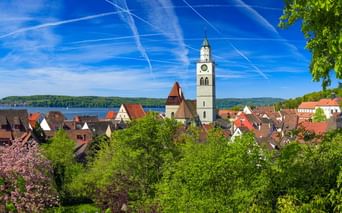 Panoramablick auf Überlingen mit weißem Kirchturm, roten Ziegeldächern und dem Bodensee im Hintergrund unter blauem Himmel.