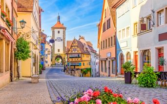 Cobblestone street in Rothenburg with colorful half-timbered houses and Siebersturm tower with orange roof. Pink and red flowers bloom in foreground.
