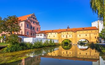 Historic Amberg Water Gate with double arches reflected in calm water. Pink Renaissance building and orange-roofed structures under blue sky.