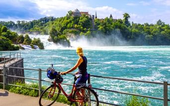 Radfahrerin mit rotem Fahrrad betrachtet den Rheinfall von einer Aussichtsplattform. Türkisfarbenes Wasser fließt an grünen bewaldeten Hügeln mit Schloss vorbei.