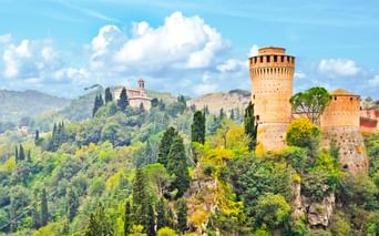 Mittelalterlicher Torre dell'Orologio-Turm auf felsigem Hügel, umgeben von Zypressen und üppiger Vegetation, mit italienischen Hügeln im Hintergrund.
