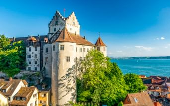 Historische Burg Meersburg mit weißen Mauern und Türmen über rotgedeckten Häusern mit Blick auf das blaue Wasser des Bodensees.