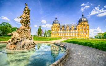 Gelbes barockes Schloss Seehof mit Kuppeltürmen hinter ornamentaler Brunnenskulptur. Kiesweg führt durch grüne Rasenflächen unter blauem Himmel.