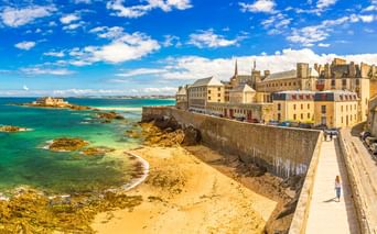Sandstrand entlang historischer Steinmauern in Saint-Malo mit türkisfarbenem Wasser, Felsen und historischen Gebäuden unter blauem Himmel.
