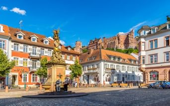Kornmarkt in Heidelberg mit historischen Gebäuden, verzierter Brunnensäule und Blick auf die Schlossruine am Hang unter blauem Himmel.