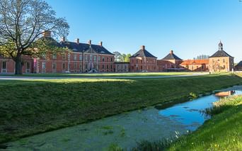 Schloss Klütz mit roten Backsteingebäuden und Wassergraben im Vordergrund. Grüne Rasenfläche und Bäume unter blauem Himmel nahe der Ostsee.