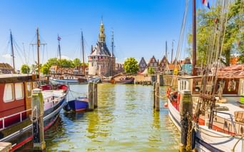 Hafenansicht in Hoorn mit traditionellen Booten an Holzpfählen. Historischer Turm und Giebelhäuser säumen das Ufer unter blauem Himmel.