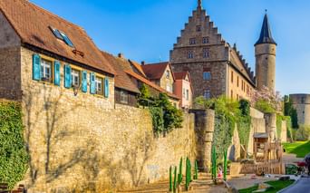 Medieval town wall in Ochsenfurt with stone houses, a stepped gable building, and a round tower. A playground sits at the wall's base under blue sky.