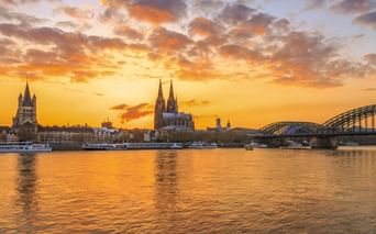 Cologne skyline at sunset with Cologne Cathedral and Hohenzollern Bridge over the Rhine. Orange and golden clouds reflect in the river water.