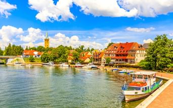 Malerisches Ufer von Mikolajki mit Booten am Ufer, bunten Gebäuden mit roten Dächern und einem Kirchturm unter blauem Himmel.