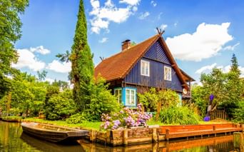 Blaues Holzhaus mit orangefarbenem Ziegeldach am Wasser im Spreewald. Holzsteg mit Blumen und Boot im Vordergrund, umgeben von üppigem Grün.