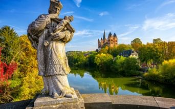 Stone statue on a bridge overlooking the Lahn River with Limburg Cathedral on a hill. Autumn trees frame the scene under a blue sky.