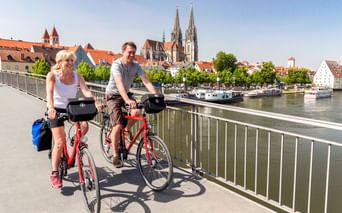 Zwei Radfahrer auf der Steinernen Brücke in Regensburg mit der Donau und historischer Altstadt-Silhouette mit Domtürmen im Hintergrund.