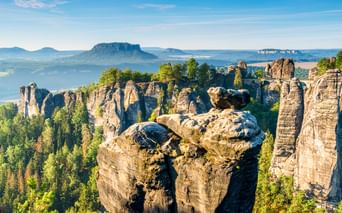 Dramatische Sandsteinfelsen der Bastei bei Rathen mit grünem Wald, fernen Tafelbergen und blauem Himmel in der Sächsischen Schweiz.