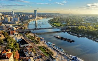 Luftaufnahme von Belgrad mit der Donau, Brücken, Kreuzfahrtschiffen und Stadtsilhouette unter blauem Himmel mit sichtbaren grünen Parks.