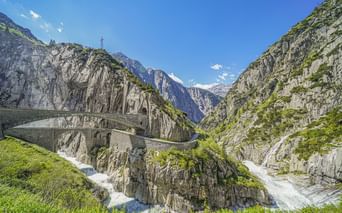 Historische Steinbrücke über die Schöllenenschlucht beim Gotthardpass. Steile Felswände rahmen einen reißenden Fluss ein, Berge im Hintergrund.
