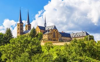 Michaelsberg Abbey in Bamberg with twin spires rising above green trees on a hilltop under a blue sky with white clouds.