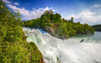 Rheinfall bei Schaffhausen mit kraftvoll herabstürzendem Wasser, üppiger grüner Vegetation und einem historischen Gebäude auf der Felsklippe.