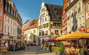 Pedestrian square in Kitzingen's old town with colorful half-timbered buildings, outdoor cafes with yellow umbrellas, and market stalls.