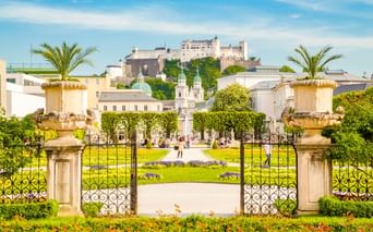 Verziertes Eingangstor mit Steinsäulen, Urnen und Palmen, das zu den Mirabellgärten mit bunten Blumenbeeten und der Festung Salzburg auf dem Hügel führt.