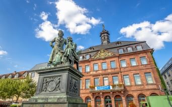 Bronzedenkmal der Brüder Grimm in Hanau mit historischem Gebäude aus rotem Sandstein dahinter. Zwei Figuren stehen auf verziertem Sockel.