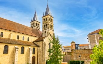 Stiftskirche Notre-Dame in Melun mit zwei romanischen Türmen mit Spitzdächern, Steinmauern und umliegenden Gebäuden unter blauem Himmel.
