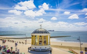 Gelb-weißer Pavillon mit Kuppel am Strand von Borkum. Sandstrand mit Besuchern, blaues Meer und bewölkter Himmel. Schiff am Horizont.