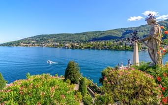 Steinstatue in einem üppigen Garten mit Blick auf das blaue Wasser des Lago Maggiore mit grünen Hügeln und einer Stadt am gegenüberliegenden Ufer.