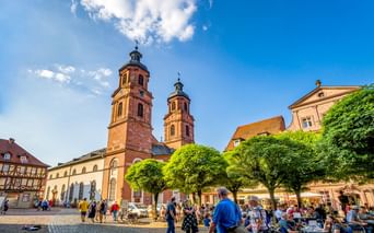 Historischer Marktplatz in Miltenberg mit Kirche mit zwei Türmen, Fachwerkhäusern und Menschen in Straßencafés unter grünen Bäumen.