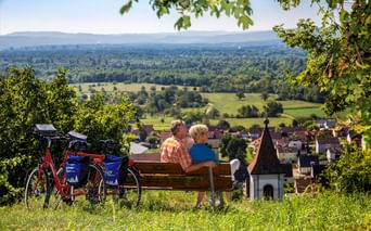 Zwei Radfahrer auf einer Bank mit Blick auf die Rheinebene bei Istein. Rote Tourenräder mit blauen Taschen stehen daneben. Dorf und grüne Felder unten.